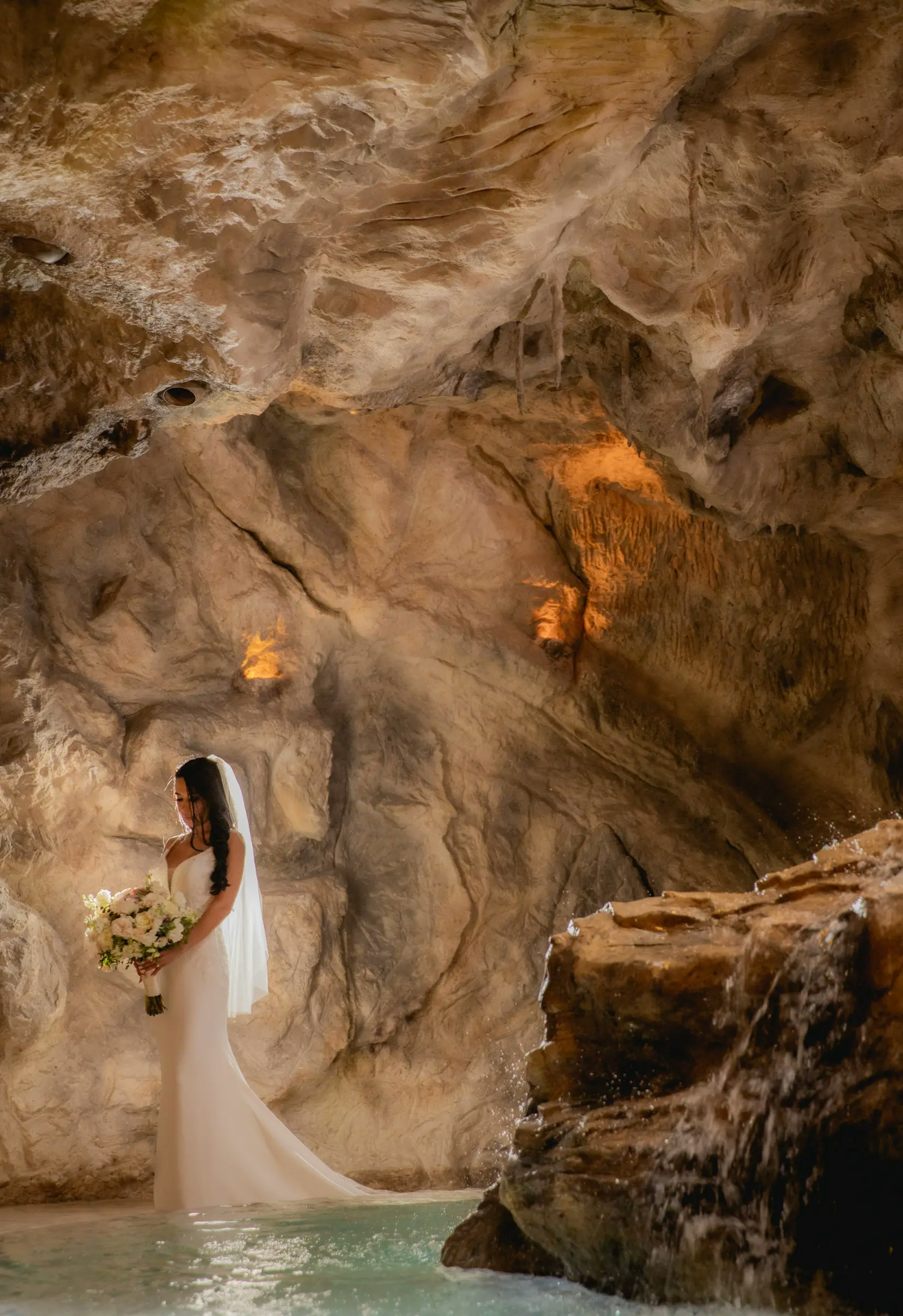 A serene bride holding a bouquet stands in a Riviera Maya cenote, her figure illuminated by the natural light that filters through the cave, enhancing the ethereal atmosphere and the natural beauty of the surroundings.