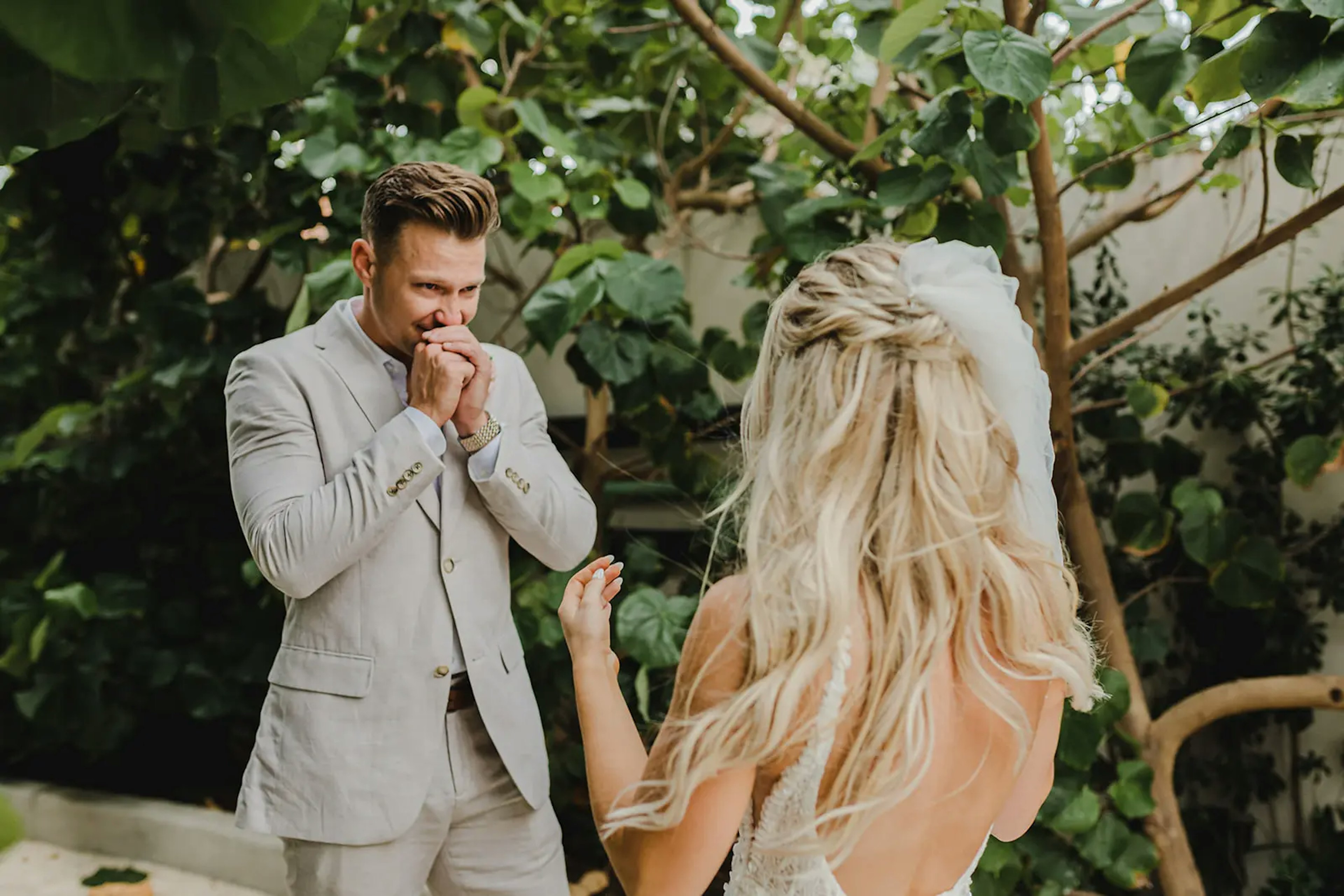 A groom, emotionally moved, holds his hands to his mouth as he gazes at his bride, who is facing away from the camera, set against a backdrop of lush greenery.