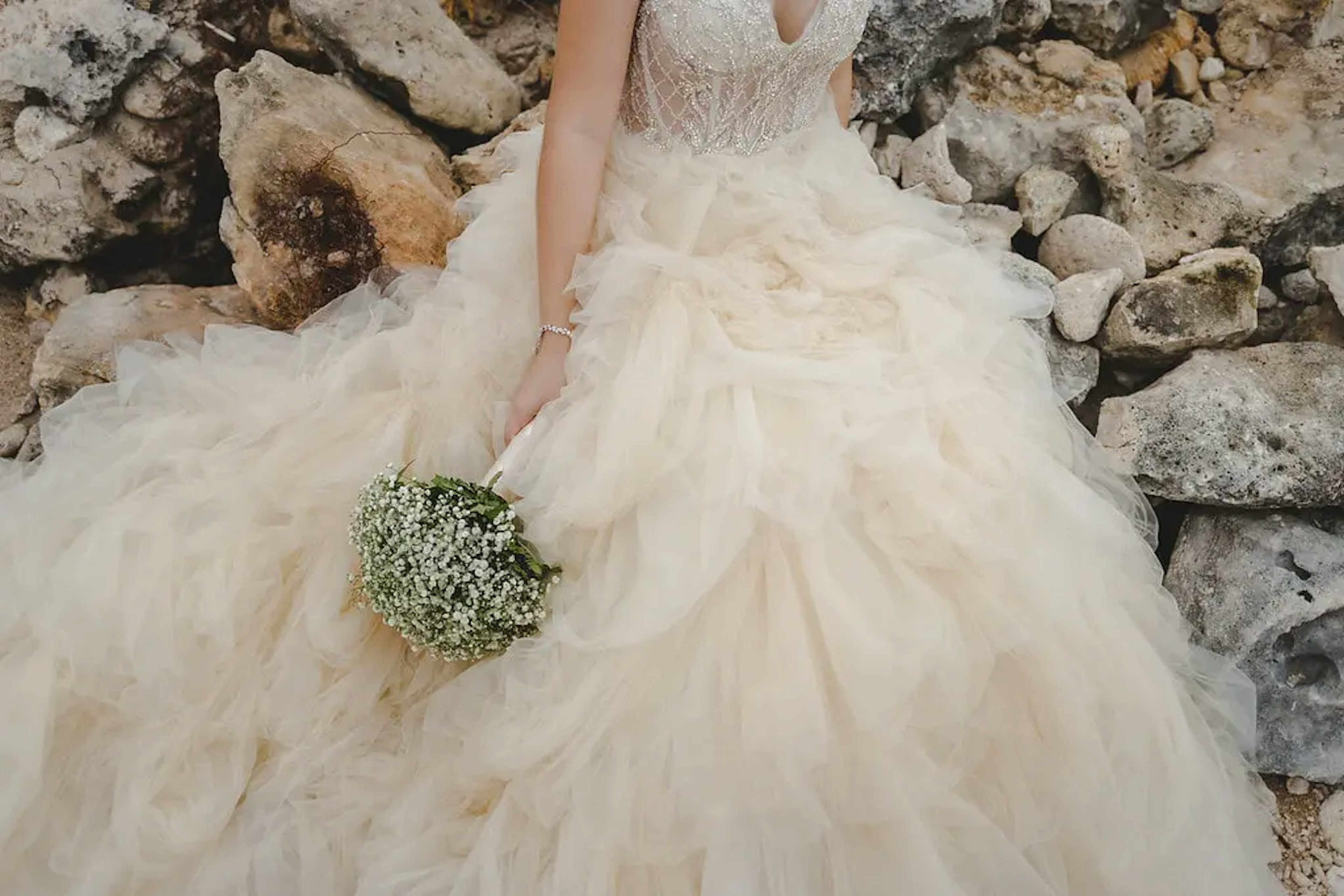 A close-up of a bride holding a delicate baby's breath bouquet, with a detailed view of her ornate lace bodice and voluminous tulle wedding gown against a rugged stone backdrop.
