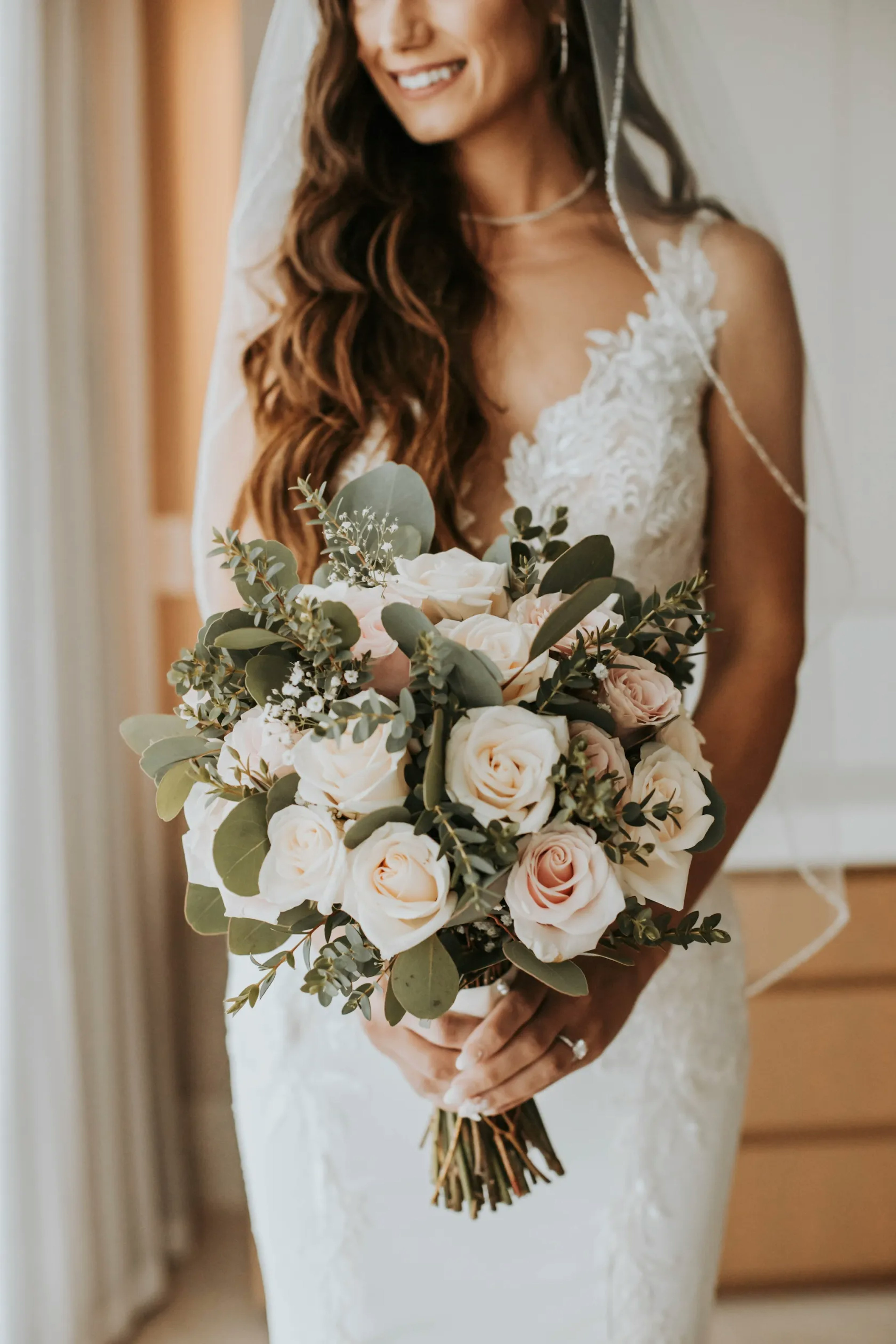 A joyful bride with a flowing veil and cascading hair holds a beautiful bouquet of pale roses and greenery, the warmth of her smile enhancing the romantic and elegant essence of the moment.