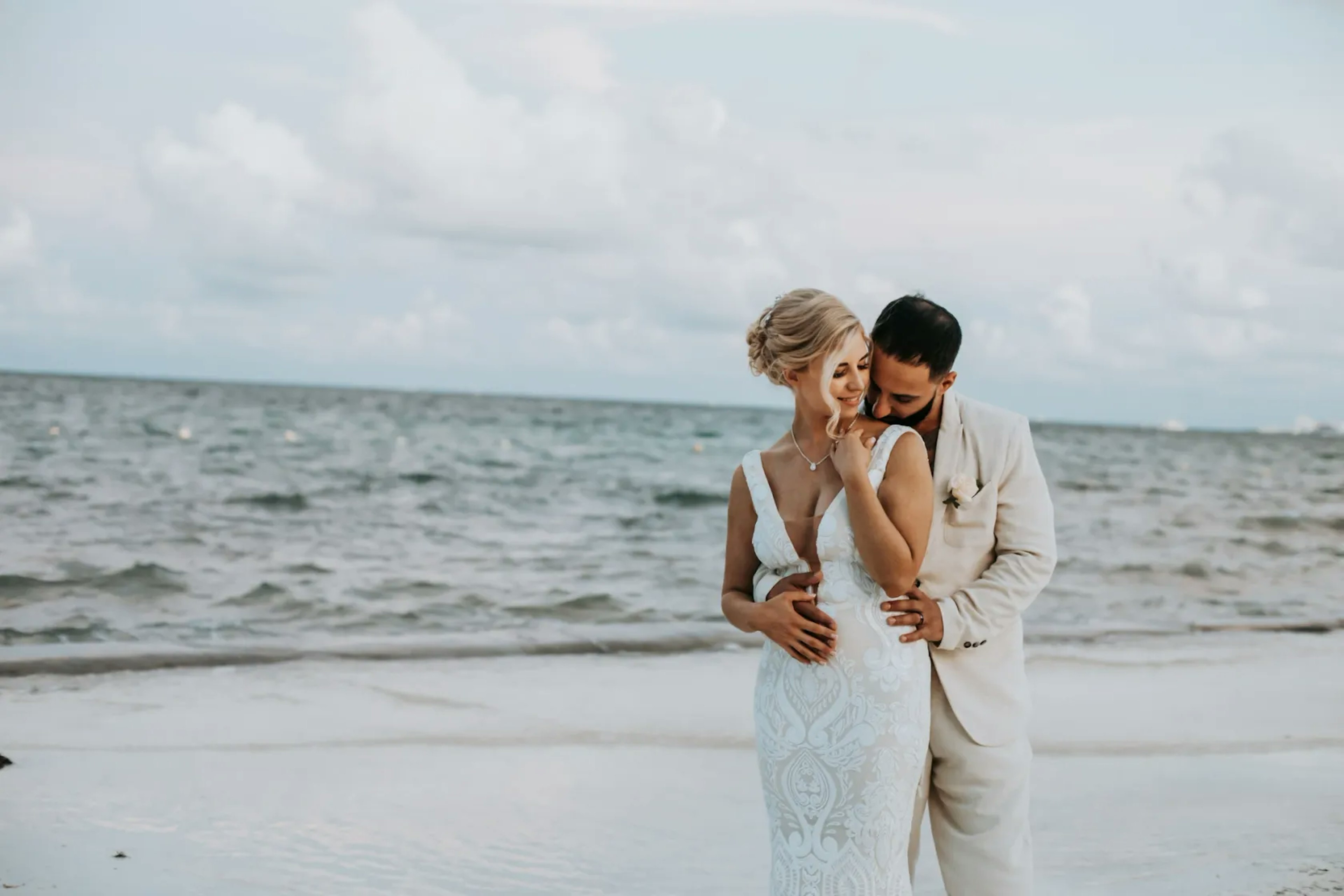 A tender moment on the beach captured between a groom, whispering to his bride, who wears a detailed lace wedding dress, both sharing a serene and intimate embrace against the backdrop of the sea.