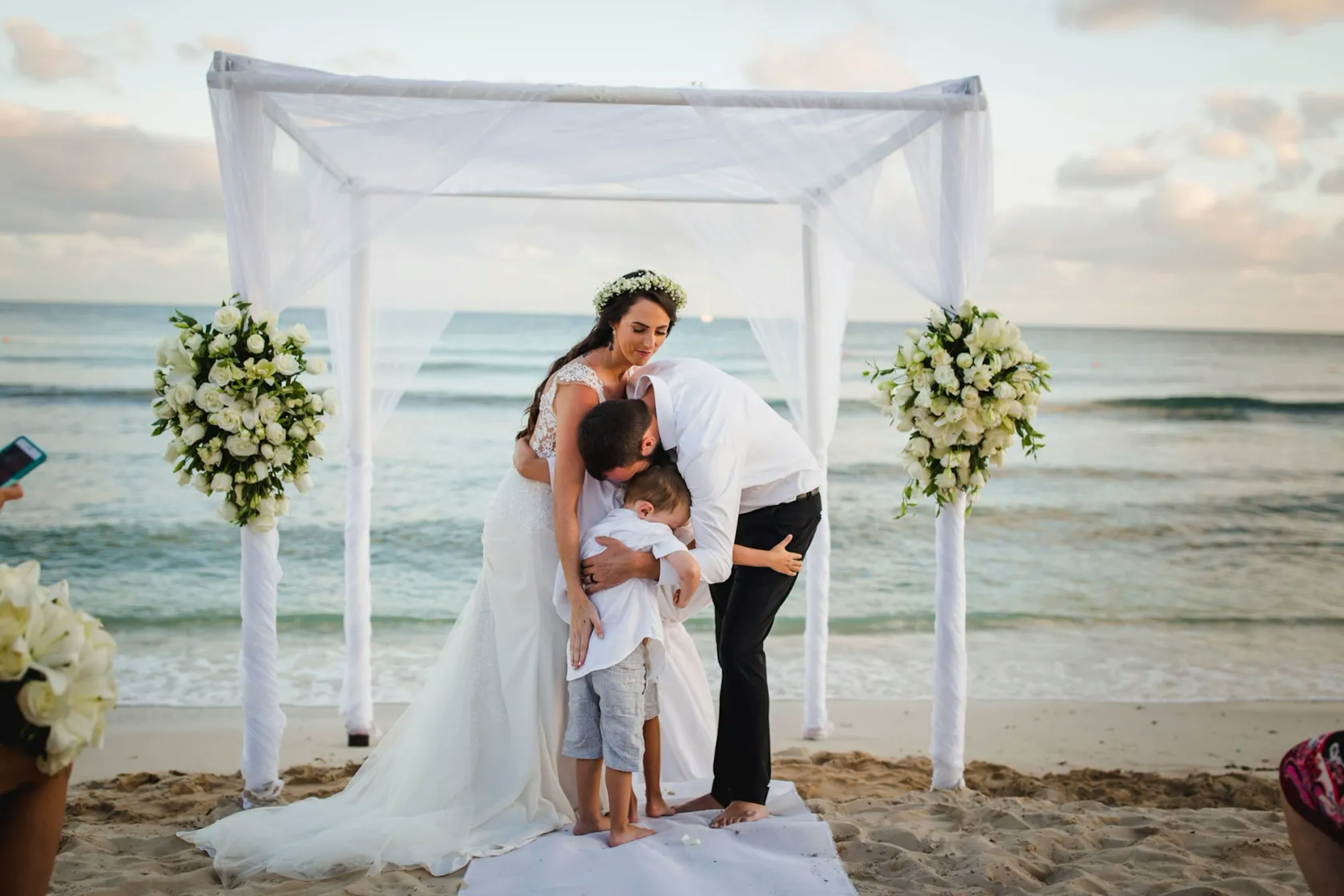 A family moment at a beach wedding: A bride with a flower crown and a groom in white embrace a young child, under a sheer white canopy adorned with flowers, with the ocean as a serene backdrop.