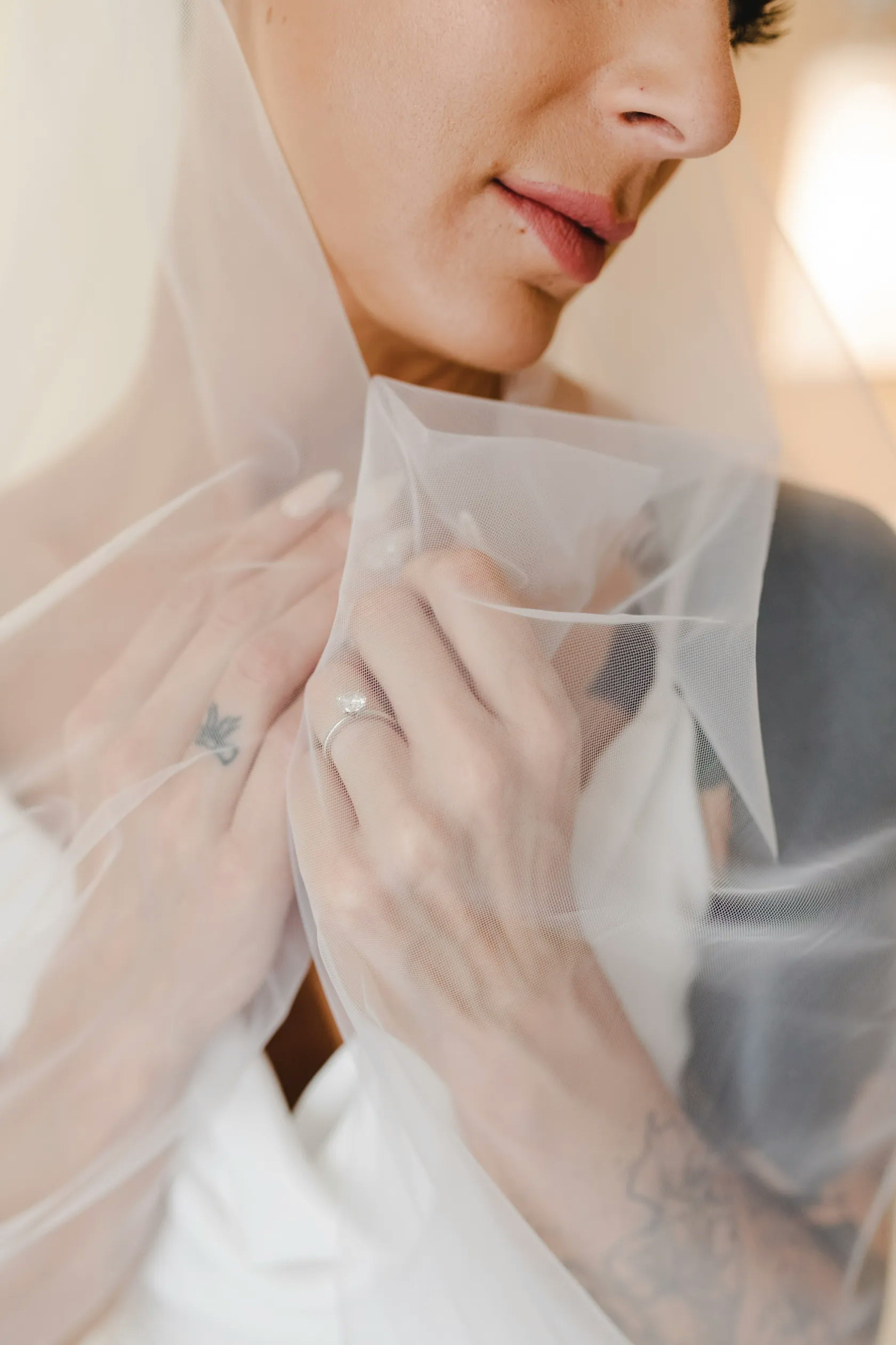 A bride in contemplation, her delicate features partly veiled, with a focus on her soft smile, a small tattoo on her shoulder, and her hand gently touching the sheer fabric of her veil.