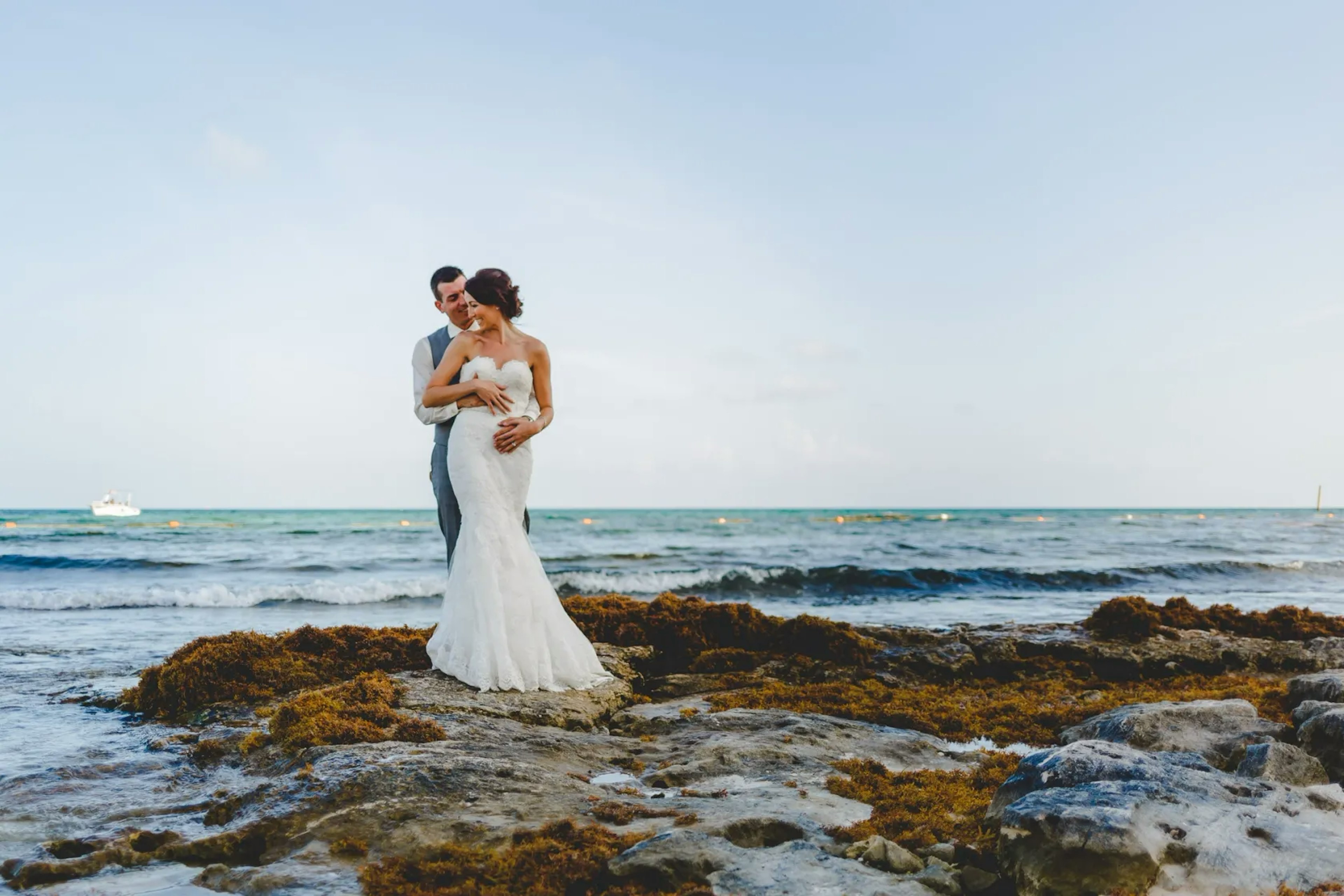 A bride and groom standing closely together on a rocky shore, with the ocean behind them and a boat visible in the distance under a clear sky.