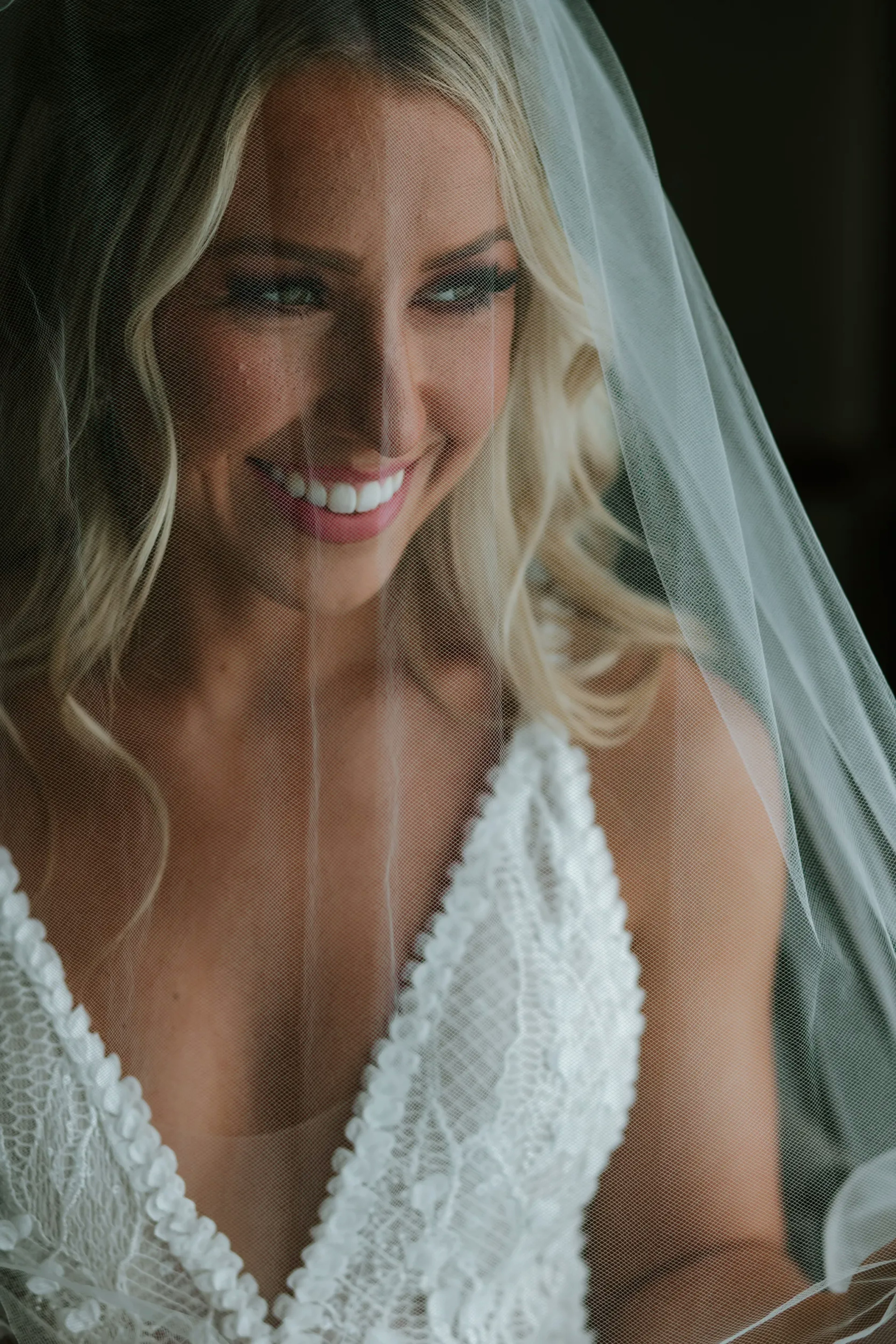 A radiant bride smiles warmly, her happiness captured through her bridal veil, with a close-up showing intricate lace details of her gown.