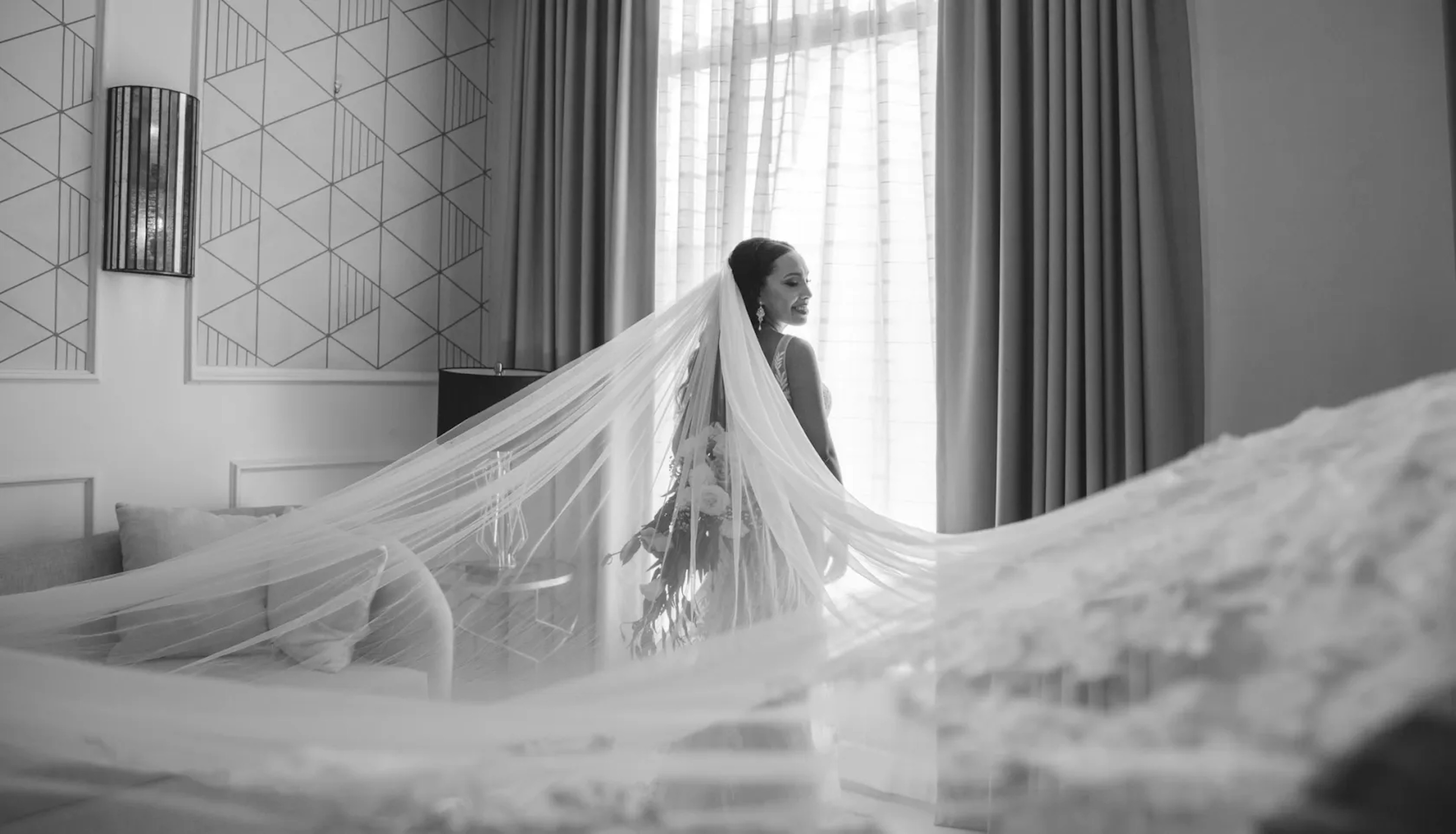 A bride stands poised in a monochrome room, her long veil flowing gracefully around her, with the light from a large window casting soft shadows and highlighting the elegant pattern of her lace dress.