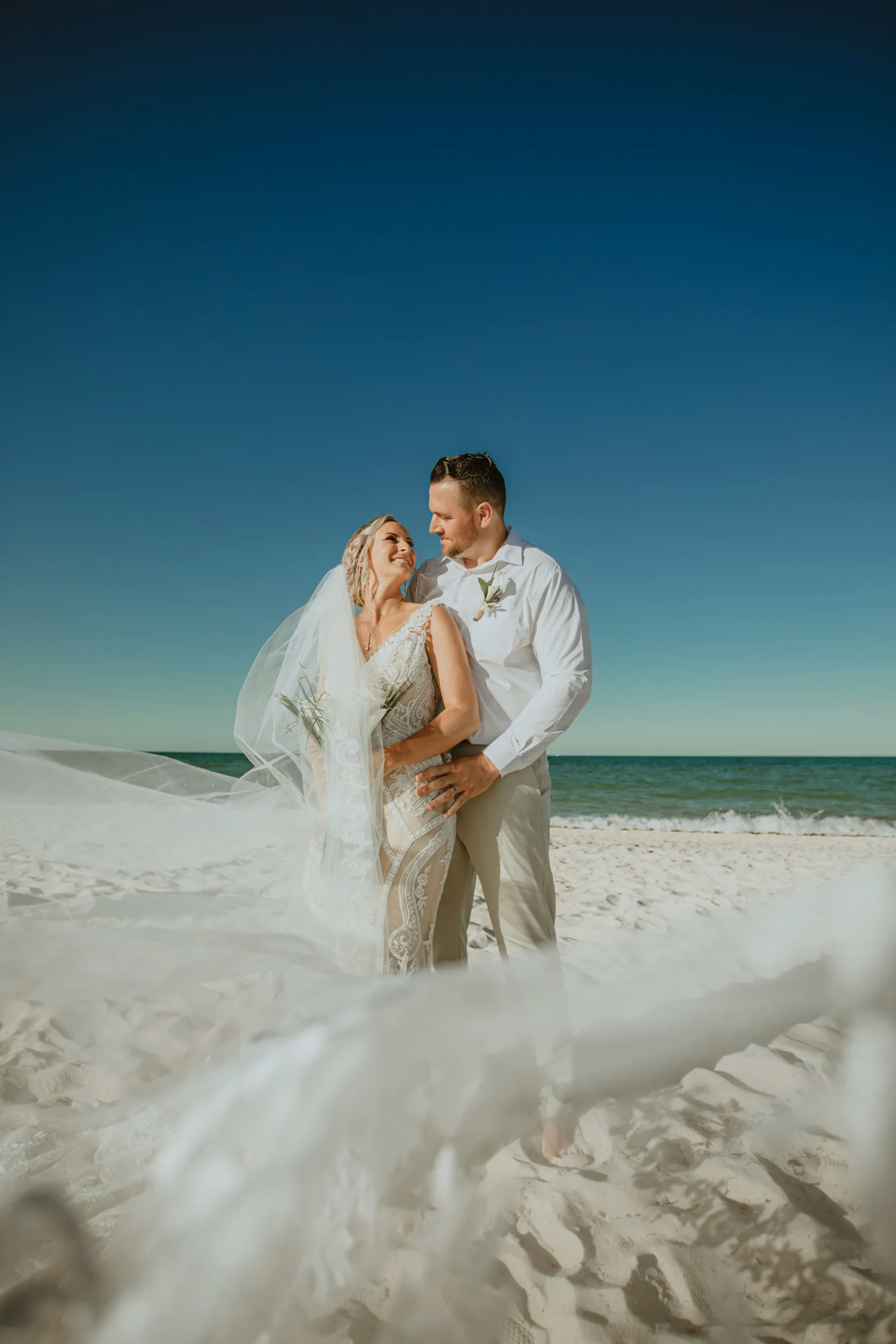 Against a clear blue sky, a bride and groom stand on a white sandy beach; the groom gazes at the bride, who smiles back, her veil caught in a gentle breeze, creating a dynamic and joyful wedding moment.