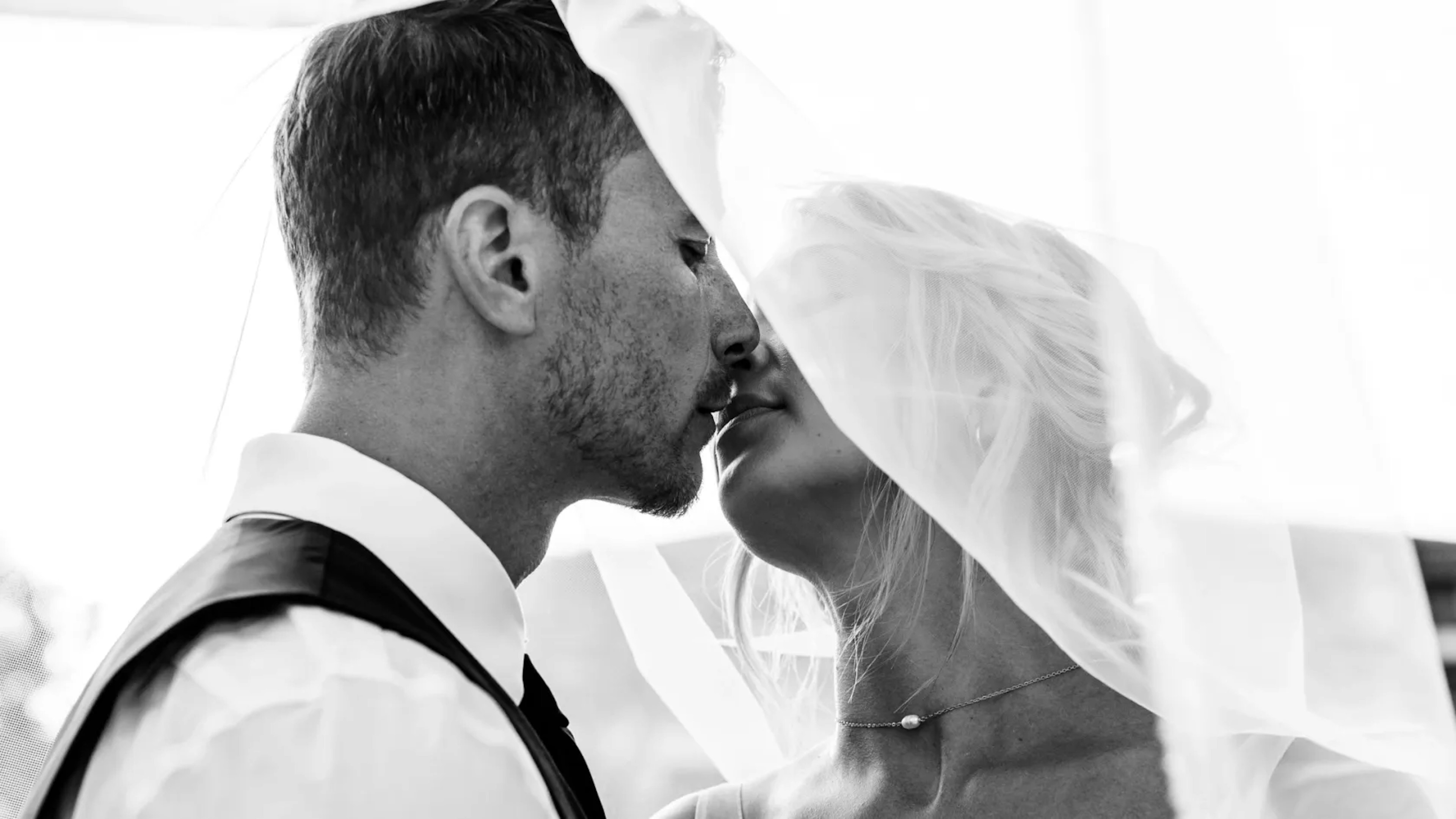 A bride and groom share an intimate moment, their faces close together, with the bride's veil softly enveloping them both in a black and white photograph that emphasizes the romantic intensity of their connection.